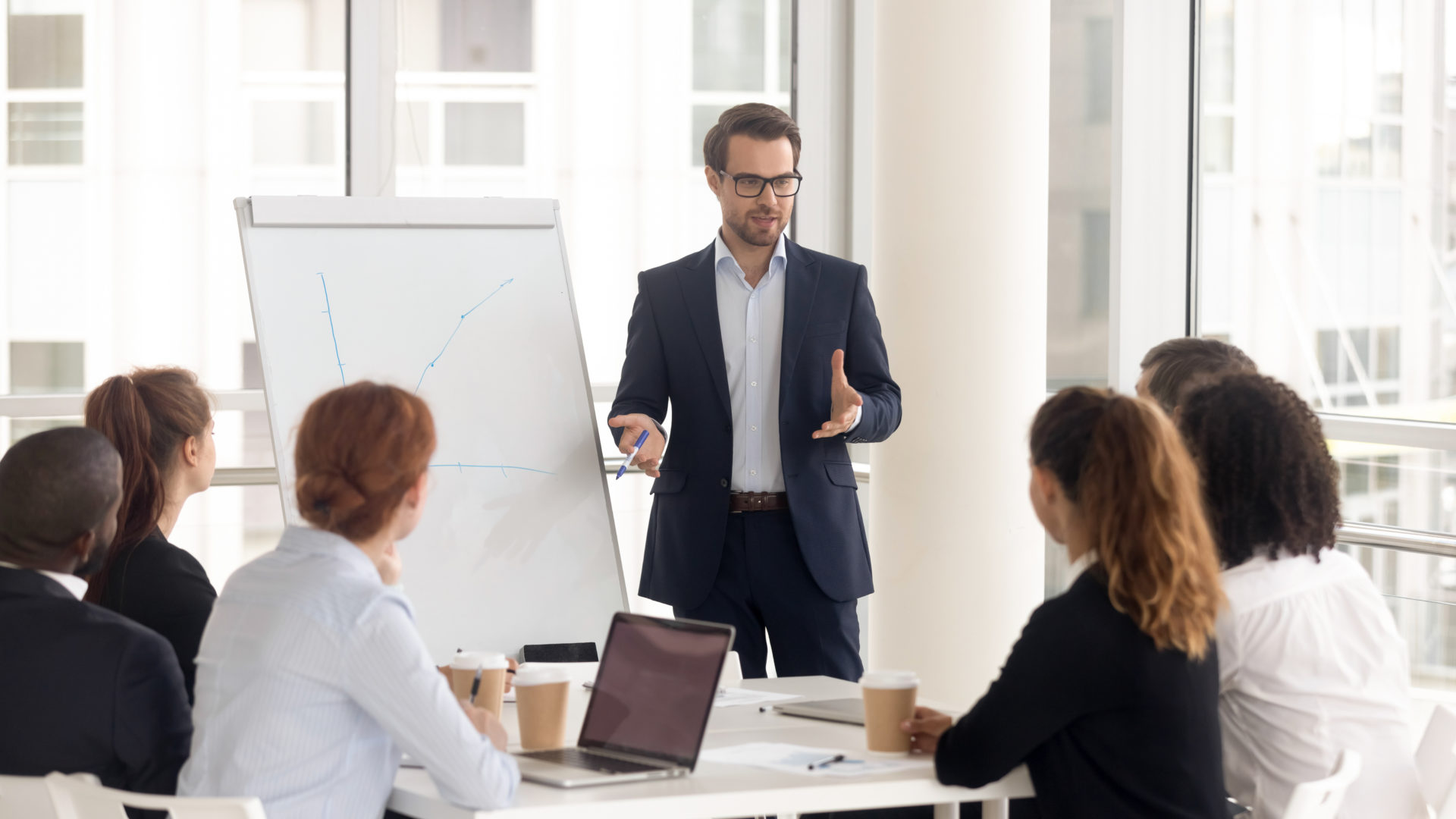 Male business coach speaker in suit give flipchart presentation, speaker presenter consulting training persuading employees client group, mentor leader explain graph strategy at team meeting workshop